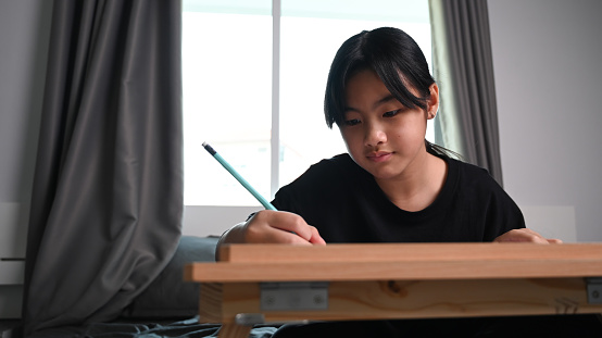 Asian child girl doing homework while sitting on bed at home.