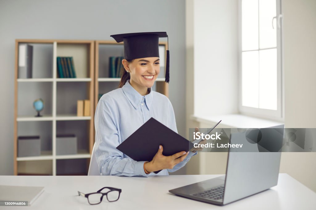 Happy graduate sitting at laptop computer and presenting her thesis remotely via video call Getting degree online. Happy student graduating from business school, college or university. Smiling woman sitting at laptop computer and presenting course work, thesis or dissertation via video call Dissertation Stock Photo