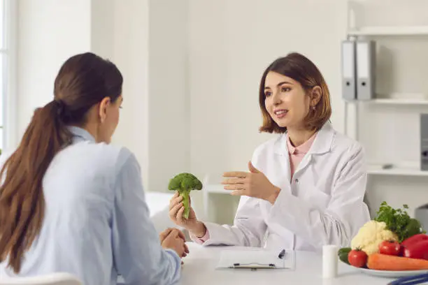 Qualified female dietitian, nutritionist, healthy food expert, doctor of alternative medicine, consultant in health center holding broccoli and telling young woman about benefits of eating vegetables