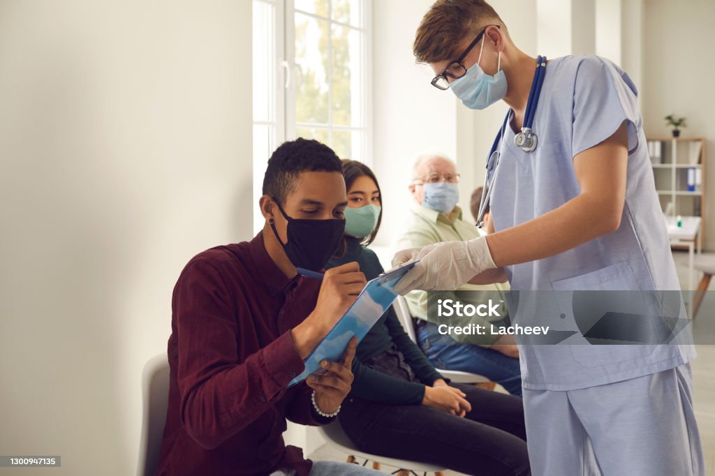 African-American man signs papers that doctor gives him before getting vaccine at the hospital Diverse people sign papers while waiting in line to get flu or Covid-19 shot at hospital, clinic or medical center. Young African-American patient putting signature on Informed Consent for Vaccination Sign Language Stock Photo