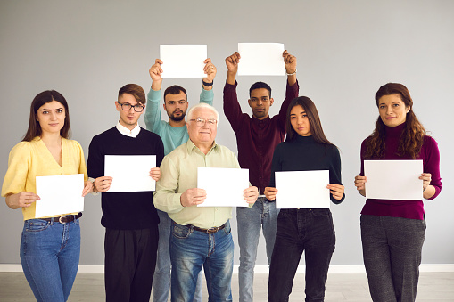 Group of different people standing together, holding blank white mockup sheets of paper and looking at camera. Team of diverse men and women holding posters with empty copy space for your message