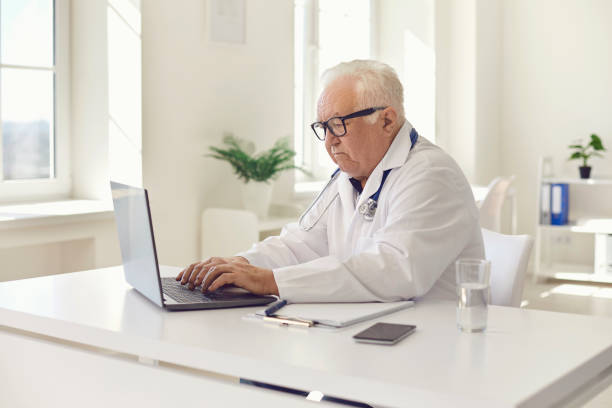 istock Serious white-haired general practitioner working on laptop sitting at desk in hospital office 1300947746