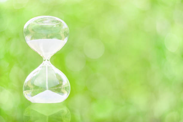 istock Time management, deadline and countdown concept : White sand fall down in a clear glass sandclock or hourglass over green bokeh space 1300948039