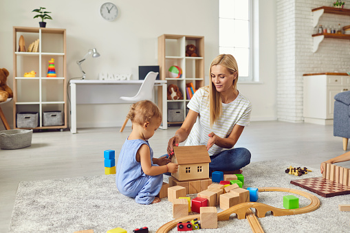 Mom and child at home. Young mother playing with little son teaching him to build toy house. Happy nanny engaging toddler boy in fun activities with wood blocks on warm floor in cozy studio apartment