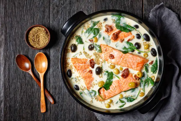 Tuscan salmon dish of pan-seared skin-on salmon fillet with cream sauce, garlic, leek, sun-dried tomatoes, olives, and spinach on a black  baking dish on a dark wooden table, top view, close-up