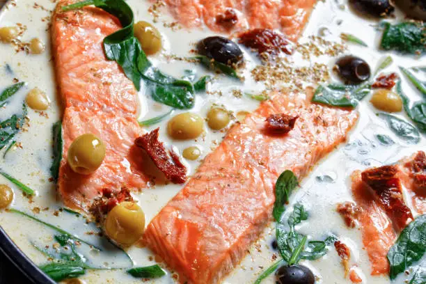 Tuscan salmon dish of pan-seared skin-on salmon fillet with cream sauce, garlic, leek, sun-dried tomatoes, olives, and spinach on a black  baking dish on a dark wooden table, top view, close-up