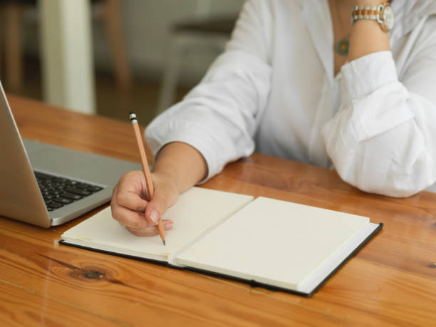 istock Female hand writing on blank notebook while working with laptop on wooden table 1300953300