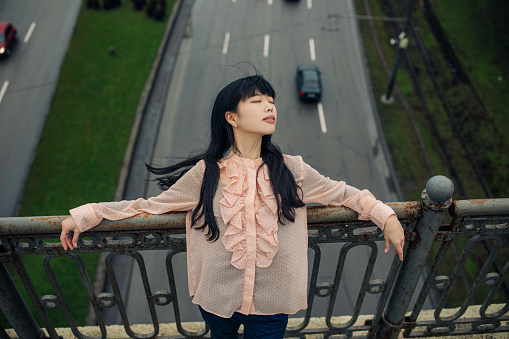 Young Japanese woman stands and relaxes on overpass against background of road and cars.