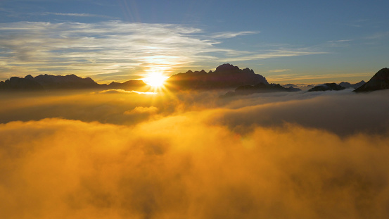 DRONE, SUN FLARE: Flying over clouds and a rocky ridge high in the French Alps at sunrise. Golden evening sun rays illuminate the mountain range and clouds gathered above the French countryside.