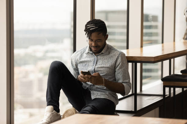 istock Young african guy sitting on panoramic window sill using smartphone 1300971861