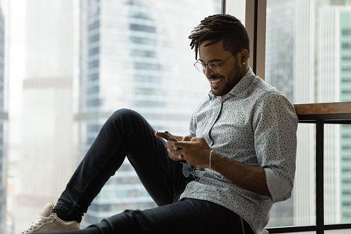 Happy millennial black man sit by big window at modern studio apartment having virtual dating flirting with beloved female by phone. Smiling african american guy spend leisure time at home using cell