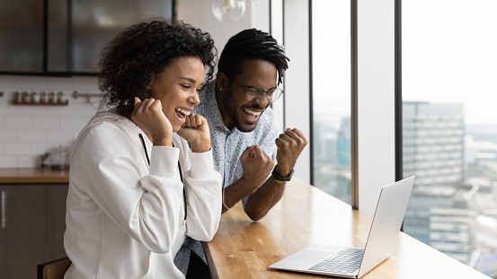 Great surprise. Excited afro american family couple scream raise hands celebrate getting mortgage loan approval from bank. Shocked young black husband wife read amazing news in email on laptop screen