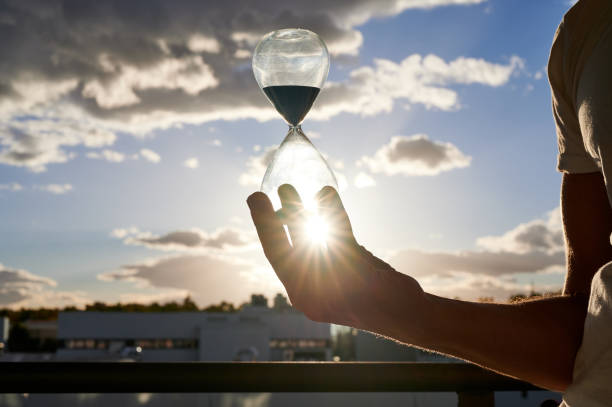 istock man's hand holding backlited hourglass with a half cloudy sky in the background 1300973088
