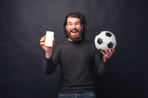 Photo of cheerful young handsome bearded man holding soccer ball and showing blank screen on smartphone.