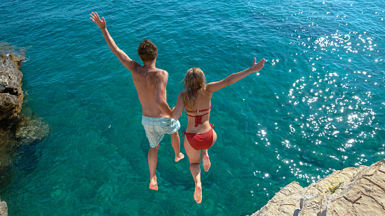 Joyful tourist couple decides to jump off a rocky cliff and dive into sea. Unrecognizable man holds his gorgeous girlfriend's hand while diving into the refreshing blue sea on a sunny summer day.