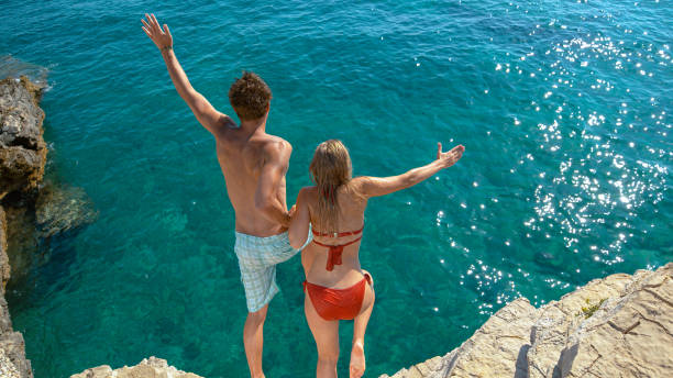 istock Young couple holding hands jumps off a cliff and into the refreshing cold ocean. 1300974498