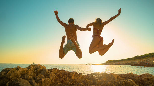 istock LOW ANGLE: Cheerful young couple holds hands while diving into ocean at sunset 1300974571
