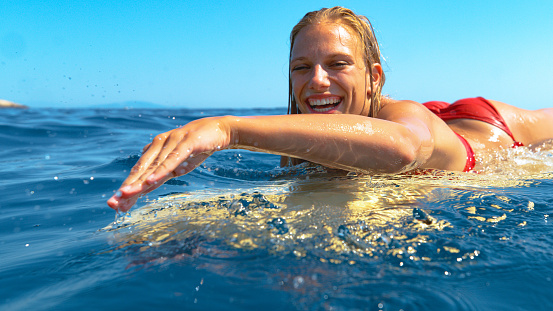 CLOSE UP, PORTRAIT, DOF: Young female tourist enjoys her summer vacation by surfing on a beautiful sunny day. Pretty blonde haired Caucasian woman smiles while swimming to line up on her surfboard.