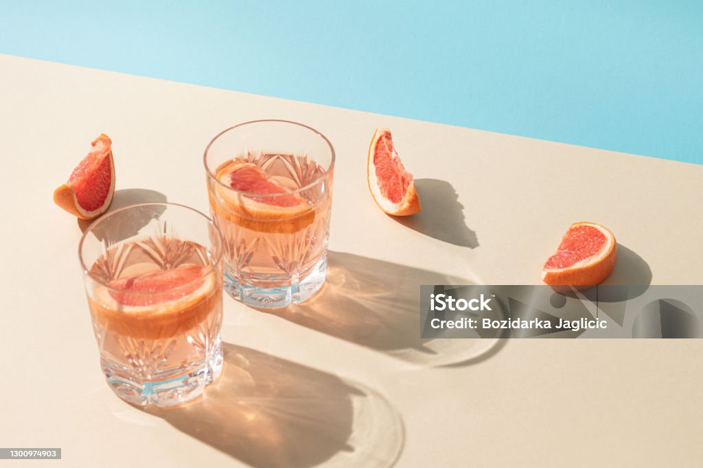 Dos copas de bebida con rodajas de pomelo fresco sobre fondo beige y azul brillante. Concepto creativo de verano minimalista. Sombras de día soleado. - Foto de stock de Cóctel - Bebida alcohólica libre de derechos