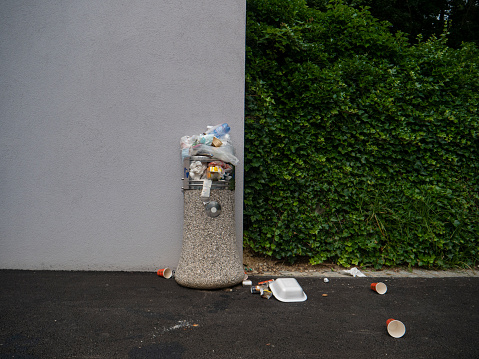 Inconsiderate people leave garbage on top and around an overflowing public bin by an empty pavement. Styrofoam food container and coffee cups lie on the ground around a waste basket full of rubbish.