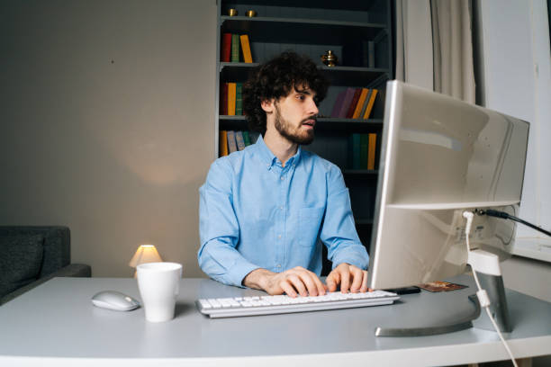 istock Side view of happy curly young man looking at camera and typing online message using computer. 1300976603