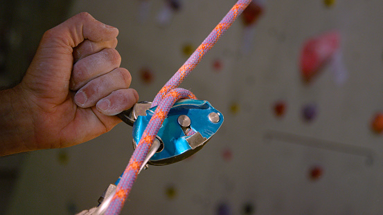 CLOSE UP, DOF: Unrecognizable belayer helps a climber descend after climbing an indoor wall. Detailed shot of a Grigri belaying device while unknown male belayer assists climber during their descent.