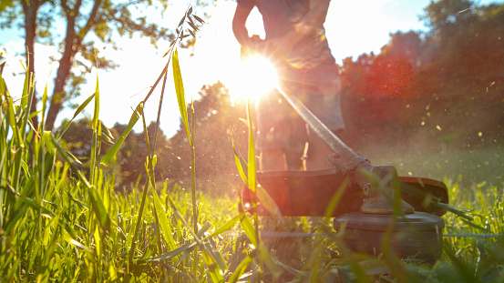 LOW ANGLE, MACRO, LENS FLARE, DOF: Young man uses a handheld grass trimmer to trim his backyard. Unrecognizable male gardener mows the lawn with a handheld lawnmower on a sunny spring afternoon.
