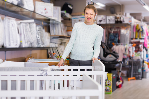 Smiling pregnant woman is choosing white crib for baby in the shop