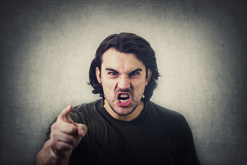 Furious young man pointing forefinger to camera blaming someone as guilty, or scolding isolated on grey wall background. Angry guy screaming and showing with index being annoyed