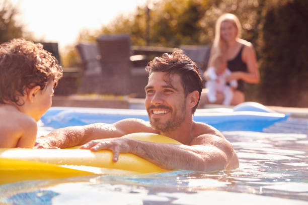 le père et le fils s’amusent à jouer dans la piscine extérieure en vacances pendant que la mère et le bébé regardent du côté - floating on water women swimming water photos et images de collection