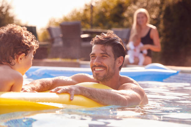 父と息子は、母親と赤ちゃんが横から見て休暇中に屋外プールで遊ぶのを楽しんでいます - floating on water women swimming pool water ストックフォトと画像
