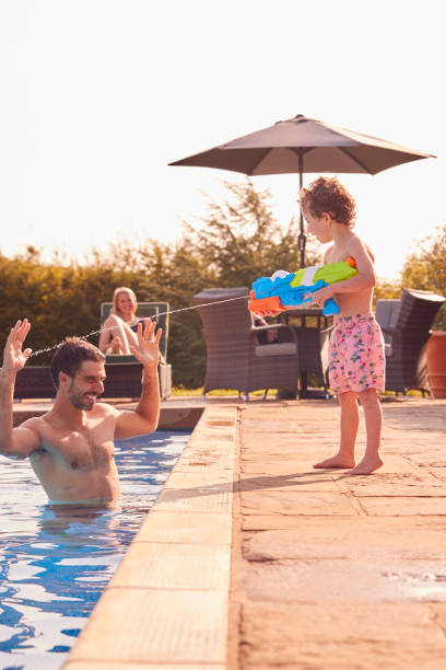 hijo squirting padre con pistola de agua jugando en la piscina en vacaciones de verano - swimming pool water people sitting fotografías e imágenes de stock