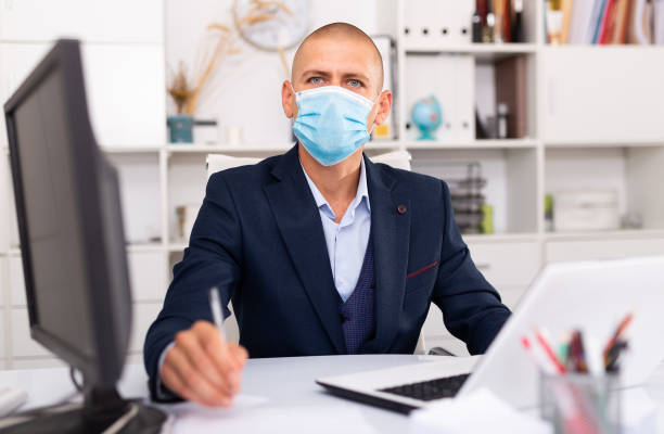 istock Professional business man in protective medical mask using laptop at workplace 1300991332