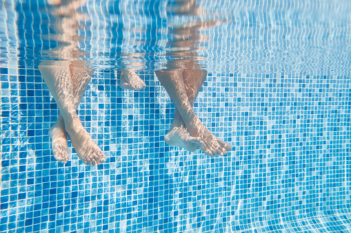 Underwater Shot Of Couples Legs As They Sit On Edge Of Swimming Pool On Vacation