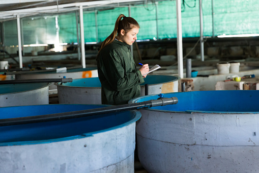 Portrait of woman inspecting tanks for rearing trout fry on farm, making notes in notebook