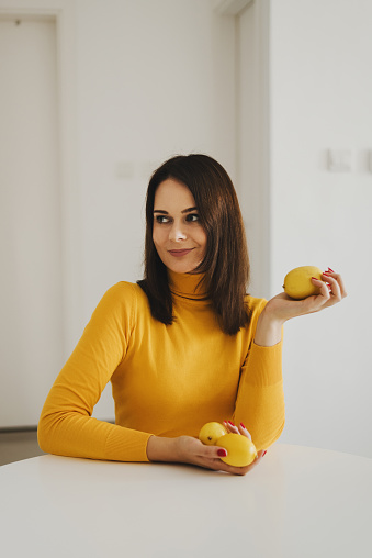 Young European woman wearing a bright yellow turtleneck sweater sitting at the dining table and holding lemons
