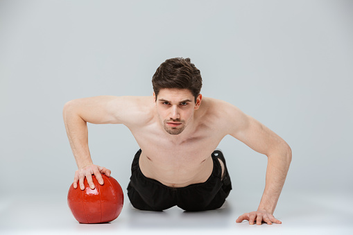 Portrait of a serious young half naked sportsman doing push ups with a heavy ball isolated over gray background