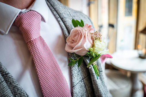 Close up of pink rose wedding favour with green leaves and stalk on grooms suit jacket with matching tie colour looking smart on day of ceremony