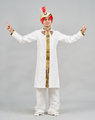 A young caucasian man is dressed in a Indian traditional clothing (Sherwani and Pheta). The male dancer is showing a poses of national dance. He is looking at the camera. Studio shooting