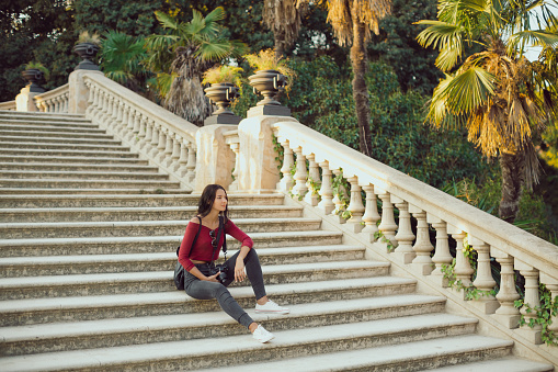 Young girl with backpack outdoors. Happy woman dressed in casual clothes sitting in a city park. Holidays and tourism concept.