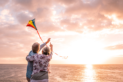 close up and portrait of two old and mature people playing and enjoying with a flaying kite at the beach with the sea at the background with sunset - active seniors having fun