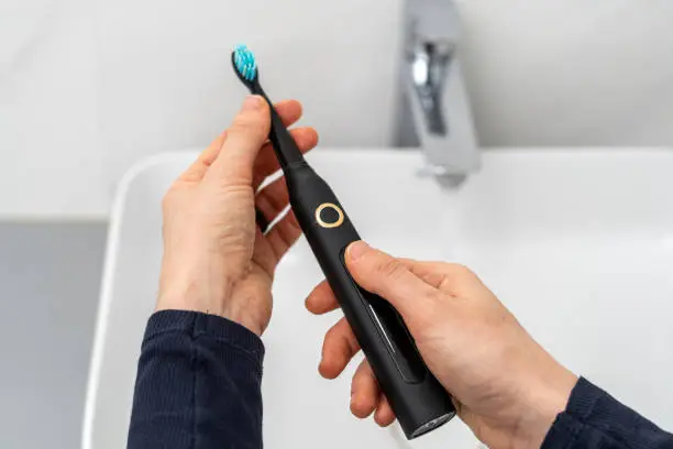 Pov woman hold professional sonic electric toothbrush with replaceable brush in hands over sink on blurred background. Wireless equipment and oral care concept