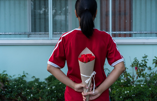 Photo of young female holding a beautiful artificial flower bouquet of red rose behind her back for giving to lover or couple on Valentine's day. Couple love, Family relationship, and LGBT Concept