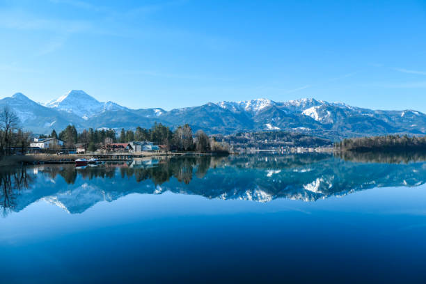 Faakersee - A panoramic view on the Faaker lake in Austrian Alps. The lake is surrounded by high mountains. Calm surface A panoramic view on the Faaker lake in Austrian Alps. The lake is surrounded by high mountains. Calm surface of the lake reflects the surrounding. Sweet flag at the shore. Clear blue sky. Serenity villach stock pictures, royalty-free photos & images
