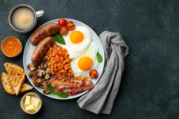 istock Full English breakfast on a plate with fried eggs, sausages, bacon, beans, toasts and coffee on dark stone background. With copy space. Top view 1301043077
