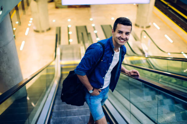 From above of smiling positive young male in casual denim clothes with sports bag going up on escalator in subway