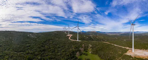 Wind farm panorama. Wind turbines and curvy road on the hill, aerial drone view. Green ecological power energy generation. Alternative energy plant, blue cloudy sky