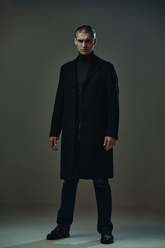 Confident serious angry guy in black coat, detective or security on gray studio background, full-length male portrait. High quality photo
