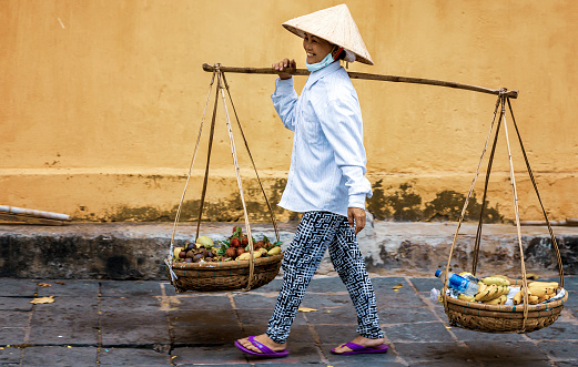 Traditionally dressed Vietnamese street vendor in Hoi-an, Vietnam carries her fruit in two baskets hanging at each end of a bamboo pole