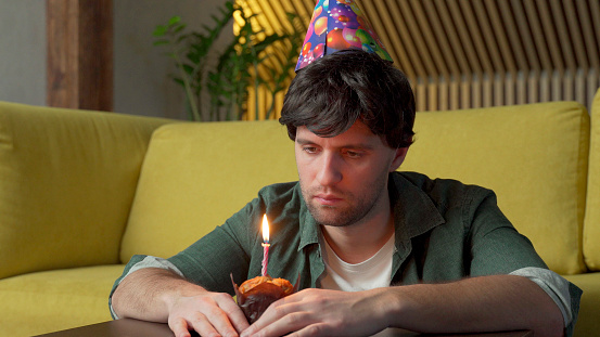 Sad man celebrates his birthday alone in the living room, blowing out the candles on the cake.